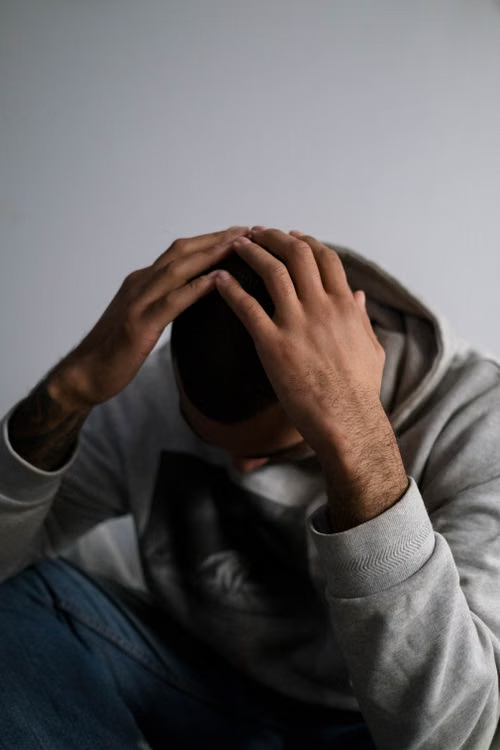  A man in a hoodie sits indoors, appearing contemplative or sad