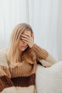 A person in a warm sweater covers her face, appearing unwell while sitting indoors.