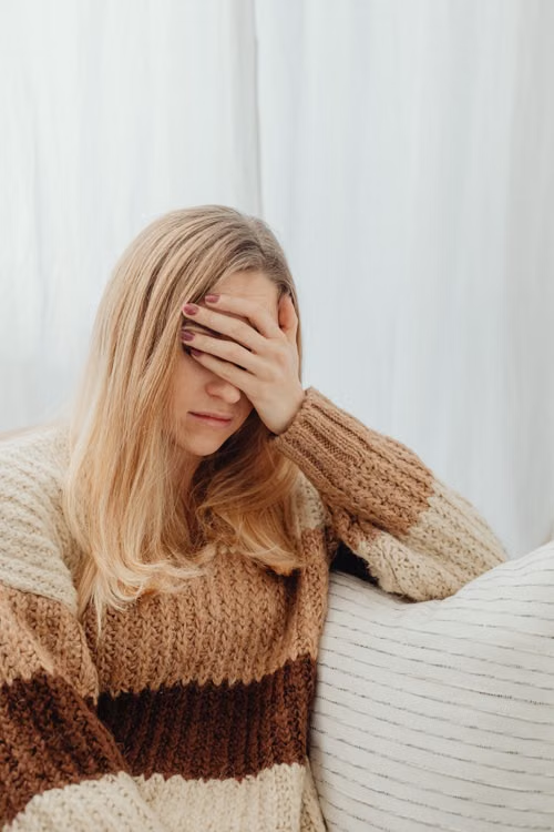 A person in a warm sweater covers her face, appearing unwell while sitting indoors.