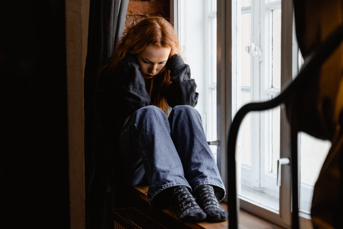 A person in a sweater sits pensively by a window.