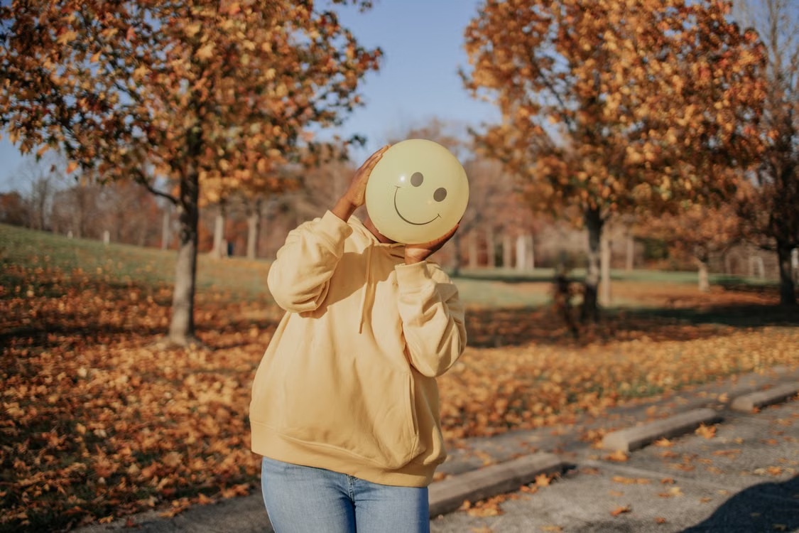 A person in a hoodie holds a smiley face balloon in a sunlit park during fall.